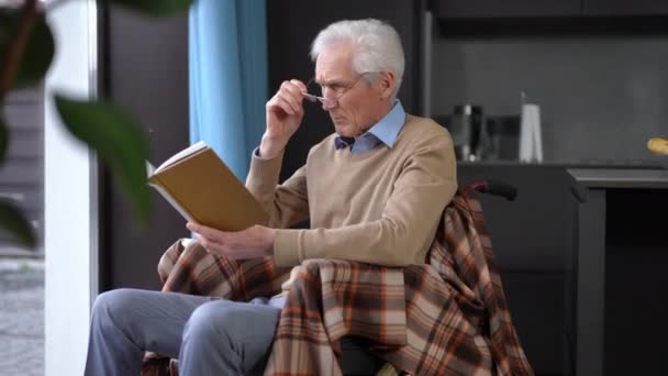坐在轮椅上 头脑清醒的老年人戴着眼镜 在室内看书 集中的白种人男性退休人员在家里享受自己的业余爱好 — 图库视频影像