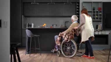 Wide shot positive Caucasian senior woman pushing wheelchair with man to countertop in kitchen. Smiling confident wife taking care of husband dining with spouse indoors talking