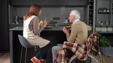 Back side view of positive senior wife dining with husband in wheelchair at home. Confident Caucasian couple of retirees enjoying food talking smiling