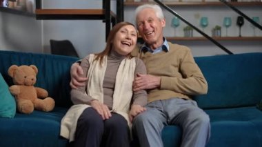 Front view portrait of relaxed senior couple hugging laughing watching comedy film on TV. Medium shot of carefree Caucasian handsome husband and beautiful wife enjoying leisure at home in living room