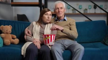Absorbed couple of retirees watching TV eating popcorn sitting on couch in living room. Front view medium shot portrait of concentrated Caucasian husband and wife enjoying movie at home indoors