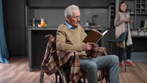 轮椅读物中聪明老人的画像 就像女人从厨房端咖啡杯一样 英俊的白人丈夫喜欢早上和妻子呆在家里的业余爱好 — 图库视频影像