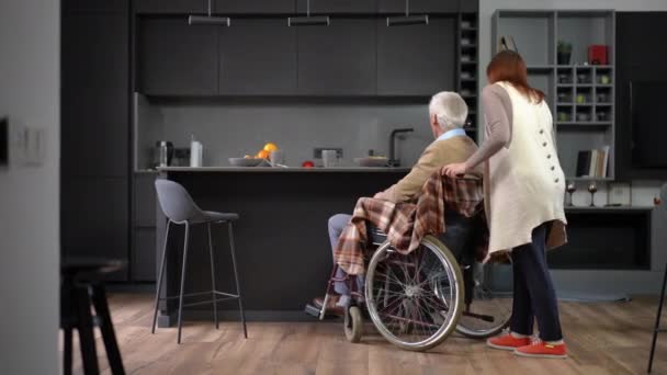 广角镜头正面白人老年妇女推着轮椅与男子在厨房的台面 带着自信的妻子和丈夫在室内吃饭聊天 — 图库视频影像