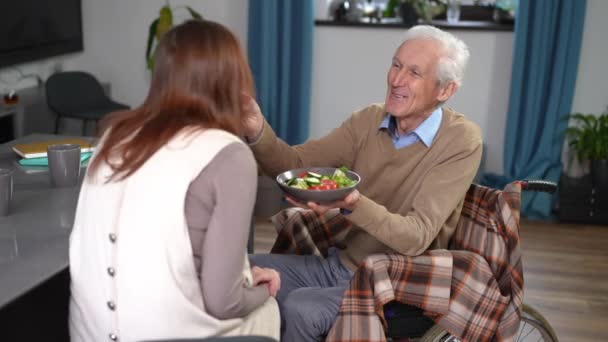 坐在轮椅上的老丈夫在家里用蔬菜沙拉喂老婆笑 笑容可亲的白人男子在室内与女人共进早餐的画像 — 图库视频影像