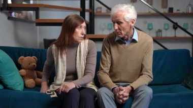 Senior Caucasian couple of retirees arguing sitting on couch in living room at home. Front view medium shot portrait of dissatisfied husband and wife quarreling indoors. Family difficulties