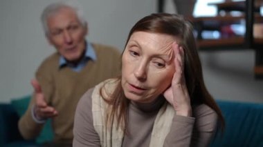 Portrait of sad Caucasian senior woman with dissatisfied facial expression listening man yelling at background. Stressed wife arguing with husband at home in living room. Irritation and conflicts