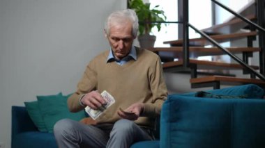 Sad lonely senior man counting cash looking away thinking sitting on couch in living room. Portrait of poor Caucasian male retiree planning budget calculating money at home