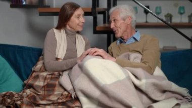 Carefree happy couple of retirees holding hands sitting on sofa talking. Smiling loving Caucasian husband and wife enjoying leisure at home in living room