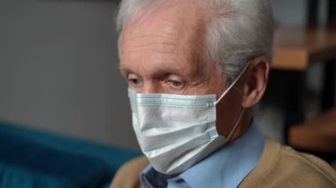 Close-up sick Caucasian senior man in Covid-19 face mask coughing wrapping in blanket looking away. Fatigue ill retiree on coronavirus quarantine with symptoms of pandemic disease