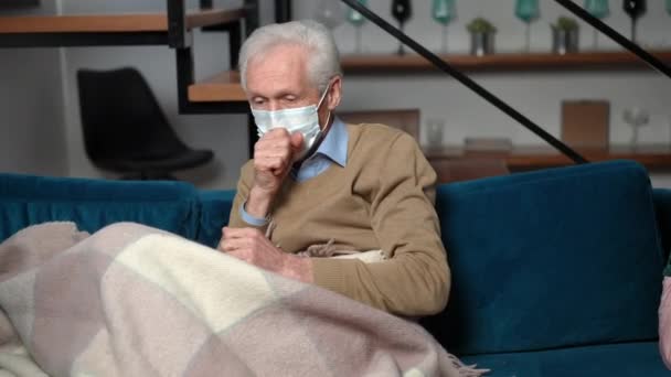 コロナウイルスの白人の先輩は リビングルームのソファの上に座って毛布に包まれたマスク咳をします 悲しい疲労の肖像病気男性退職者自宅で室内Covid 19パンデミック — ストック動画