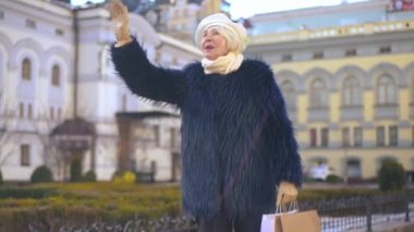 Zoom in to smiling senior elegant woman waving looking away standing on city street and looking at camera. Positive charming Caucasian retiree greeting and posing outdoors on sunny spring autumn day