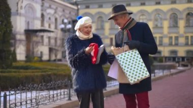 Elegant senior couple of retirees talking standing on city street with shopping bags. Positive Caucasian husband and wife with new berets smiling enjoying Black Friday sales. Family and consumerism
