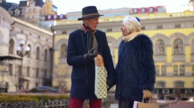 Loving senior couple of retirees holding hands walking with shopping bags in city talking. Portrait of elegant handsome Caucasian man and beautiful woman strolling on the street chatting