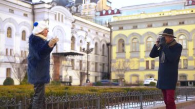 Elegant stylish senior woman posing on city street as man taking photo with camera. Positive carefree couple of Caucasian retirees enjoying tourism having fun on vacations
