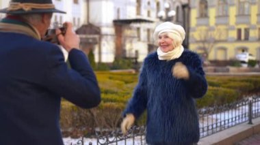 Joyful positive senior woman posing for man taking photos on city street. Smiling carefree Caucasian elegant wife enjoying travelling with husband on retirement in Europe