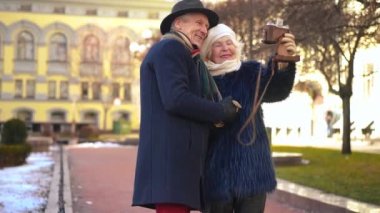 Smiling positive couple of retirees taking selfie with retro camera standing on city street. Happy relaxed Caucasian man and woman posing outdoors photographing on trip in Europe
