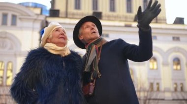 Couple of relaxed senior tourists standing on city street talking pointing looking away. Happy relaxed Caucasian man and woman chatting travelling in Europe outdoors. Retirement and tourism