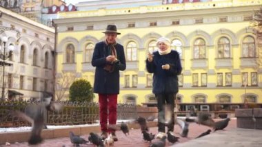Wide shot positive couple of retirees feeding pigeons on city street talking smiling. Happy confident Caucasian elegant husband and wife enjoying leisure outdoors in European town