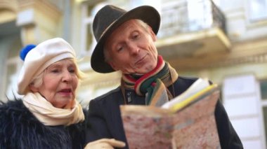 Positive senior Caucasian couple talking choosing route travelling outdoors. Portrait of confident elegant husband and wife standing with paper map on city street thinking chatting