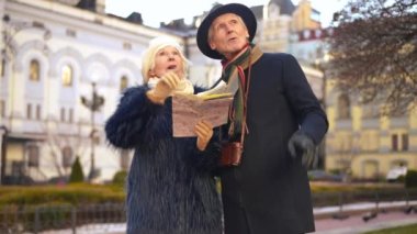 Excited curios couple of retirees with paper map pointing up smiling leaving together. Front view portrait of happy confident Caucasian husband and wife travelling in Europe outdoors