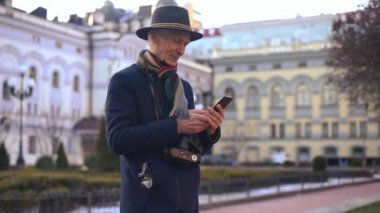 Portrait of curios senior Caucasian elegant man taking photo with smartphone app standing on city street. Smiling handsome tourist retiree enjoying journey in Europe outdoors