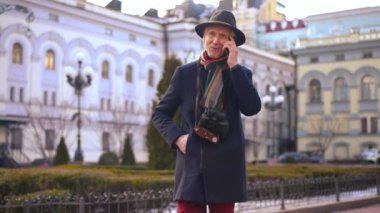 Elegant male senior tourist talking on phone walking on city street leaving. Portrait of handsome Caucasian retiree enjoying trip strolling outdoors smiling