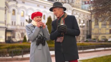 Smiling relaxed senior man and woman talking standing with tea mugs on city street. Positive happy Caucasian couple of tourists enjoying trip outdoors on autumn spring day