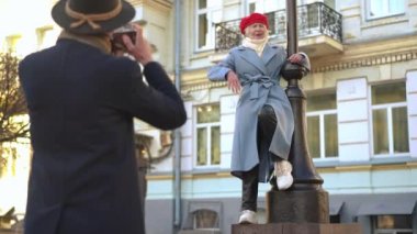 Gorgeous senior wife posing at lamp pole as husband taking photo with camera on city street. Shooting over shoulder of loving Caucasian man enjoying trip with woman in European town outdoors