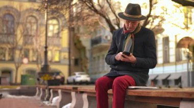 Elegant thoughtful senior man sitting on bench on the right messaging in smartphone app looking away. Portrait of handsome stylish Caucasian male retiree surfing Internet on phone on city street