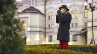 Side view senior man in Covid-19 face mask taking photo with camera standing on city street. Confident Caucasian retiree travelling in Europe on coronavirus pandemic. New normal tourism
