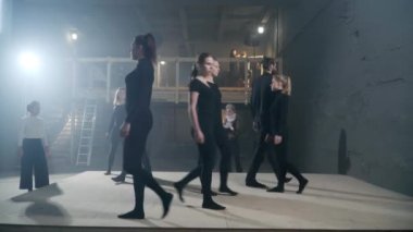 Wide shot professional men and women in black walking on stage rehearsing theatrical performance with stage director watching. Caucasian actors and actresses with serious facial expression strolling