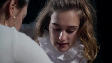 Portrait of charming young Caucasian woman in white neck ruff talking putting on stage costume with colleague helping. Close-up beautiful talented confident actress getting ready for show in theater