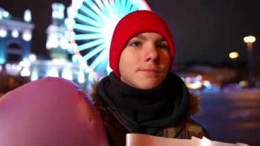 Close-up confident handsome teen boy waiting for a date on Valentines night on city street. Smiling positive Caucasian teenager with bouquet of flowers and balloon looking away. Slow motion