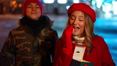 Teen Caucasian girl posing eating delicious sweets as blurred boy smiling standing at background. Positive teenage couple dating in night city on Valentines looking at camera