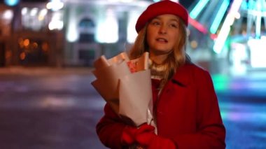 Medium shot portrait of teenage charming girl with bouquet standing in night city talking looking around. Beautiful elegant Caucasian teenager in red on Valentines outdoors. Romance and lifestyle