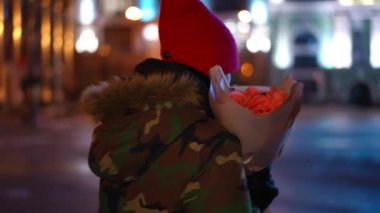 Tracking shot side view of teenage boy walking for a date with bouquet of flowers in night city. Confident Caucasian teenager with roses strolling outdoors on street. Lifestyle and romance concept