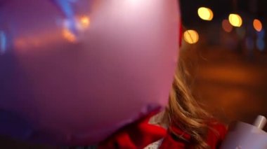 Front view close-up of joyful teen girl hiding behind heart-shaped balloon appearing grimacing posing on night city street. Portrait of cheerful happy Caucasian teenager making faces looking at camera