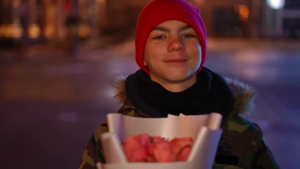 自信的白种人少年眨了眨眼 看着摄像机伸展着一束玫瑰 情人节夜晚 快乐而自信的青少年带着鲜花在城市街道上摆姿势的正面肖像 — 图库视频影像