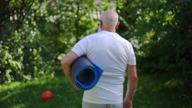 Back view old grey-haired man walking with rolled exercise mat outdoors turning looking at camera. Tracking shot of motivated Caucasian senior sportsman posing on summer day on backyard. Slow motion