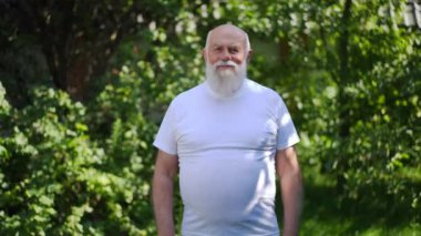 Positive confident sportive senior man gesturing strength in slow motion smiling looking at camera. Portrait of inspired old Caucasian retiree posing outdoors in park training on summer day