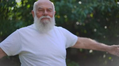 Side view motivated old grey-haired retiree with long beard exercising in slow motion turning body. Inspired Caucasian senior man working out outdoors in sunshine. Sport and healthy lifestyle