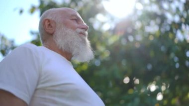 Bottom angle view of bearded confident senior man standing in sunrays outdoors on summer day. Relaxed happy Caucasian male retiree enjoying leisure in sunny park. Slow motion
