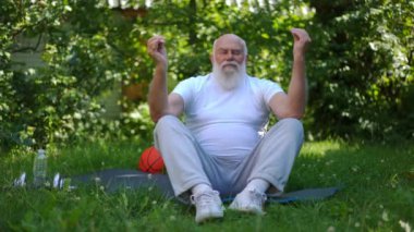 Front view portrait of confident senior Caucasian man sitting in lotus pose closing eyes. Motivated old senior retiree meditating in sunny summer park exercising. Yoga and sport concept