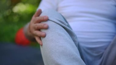 Close-up male leg with wrinkled hands rubbing knee outdoors. Unrecognizable Caucasian old sportsman stroking painful injured joint sitting in summer park. Training and aging concept