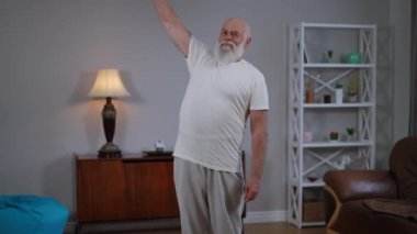Zoom in to motivated confident old bearded man lifting dumbbell looking at camera. Portrait of inspired senior Caucasian retiree posing working out training at home in living room. Slow motion