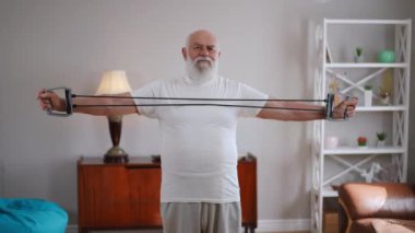 Concentrated motivated senior man stretching resistance band working out at home indoors. Medium shot portrait of confident Caucasian positive sportsman training muscles standing in living room
