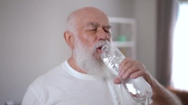 Confident old man drinking refreshing water after home workout indoors. Portrait of motivated Caucasian senior retiree after training in living room. Sport and motivation concept