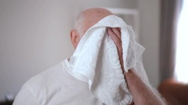 Perspiring senior sportsman rubbing face with white towel looking away smiling standing indoors. Portrait of confident satisfied Caucasian old bearded man training at home wiping sweat off forehead