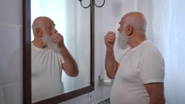 Side view old Caucasian man combing eyebrows and beard looking in mirror in bathroom. Senior male retiree getting ready at home in the morning. Domestic lifestyle and aging concept