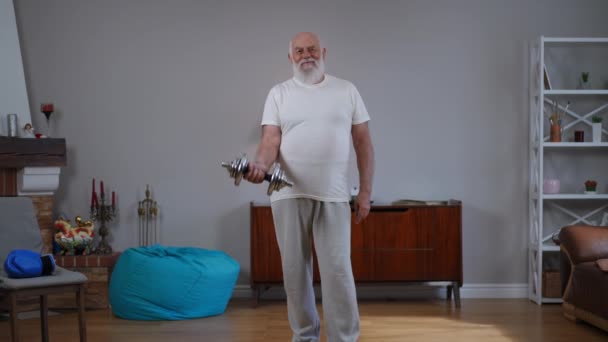 広いショットハンサムなシニア動機のスポーツマンは自宅で屋内に立ってカメラを見てスローモーションでダンベルを持ち上げる やる気を起こさせる白人の老人の肖像 — ストック動画